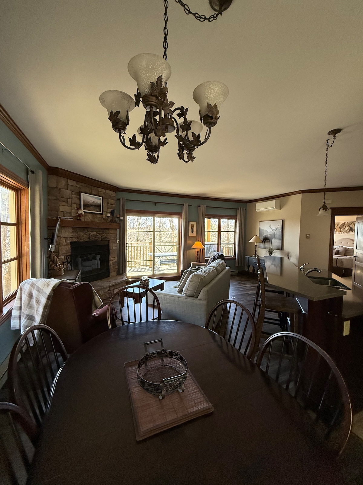 An open-concept living area is highlighted by a large dining table surrounded by wooden chairs. A chandelier hangs above, and light streams through multiple windows, illuminating a cozy seating area with a stone fireplace. A glimpse of nature is visible outside.