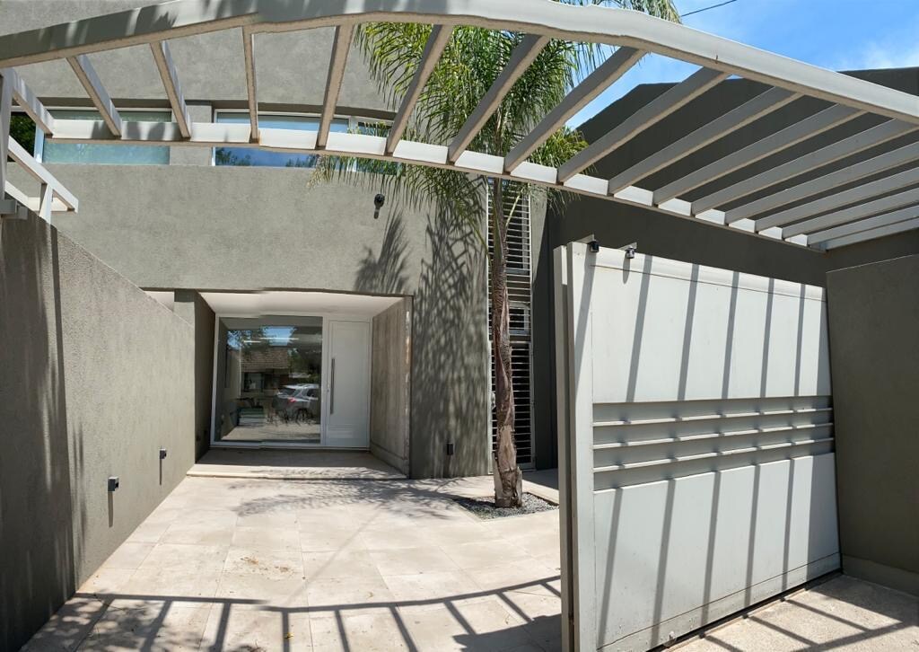 An entrance is framed by a semi-covered walkway with a modern design. A palm tree is visible to the side, and spacious glass doors lead into the interior, surrounded by smooth stone flooring, providing ease of access.