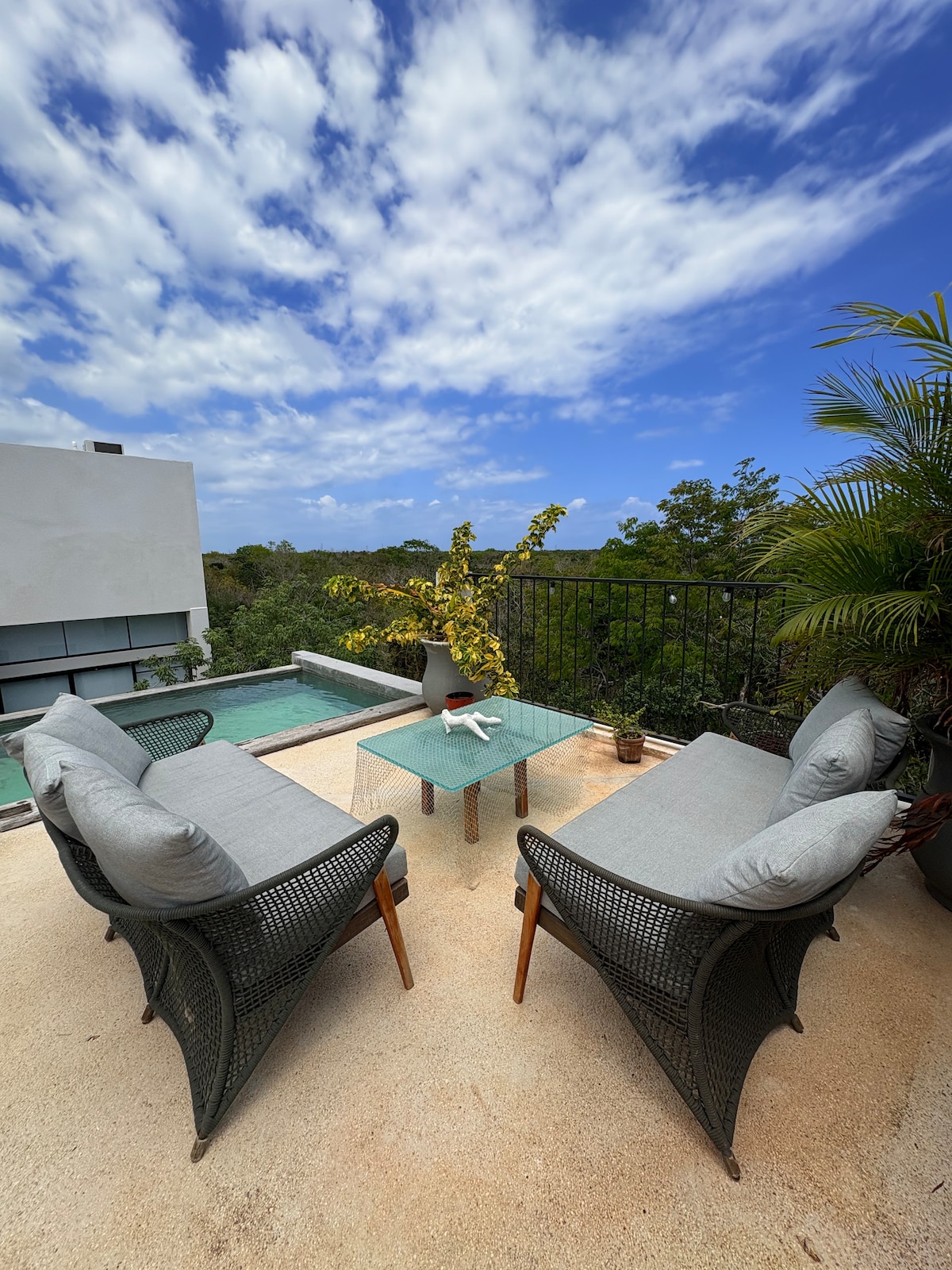 An outdoor seating area features two comfortable chairs with soft cushions arranged around a glass-top table. Lush greenery and a private pool are visible in the background, with a clear blue sky overhead, creating a serene atmosphere for relaxation.