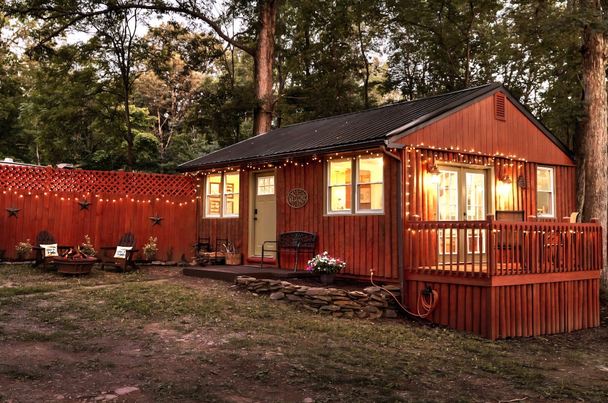 A cozy cabin exterior is showcased, featuring rich wooden walls and a welcoming front deck. String lights illuminate the space, enhancing its charm. A fire pit area is visible nearby, surrounded by seating, inviting relaxation in the natural surroundings.