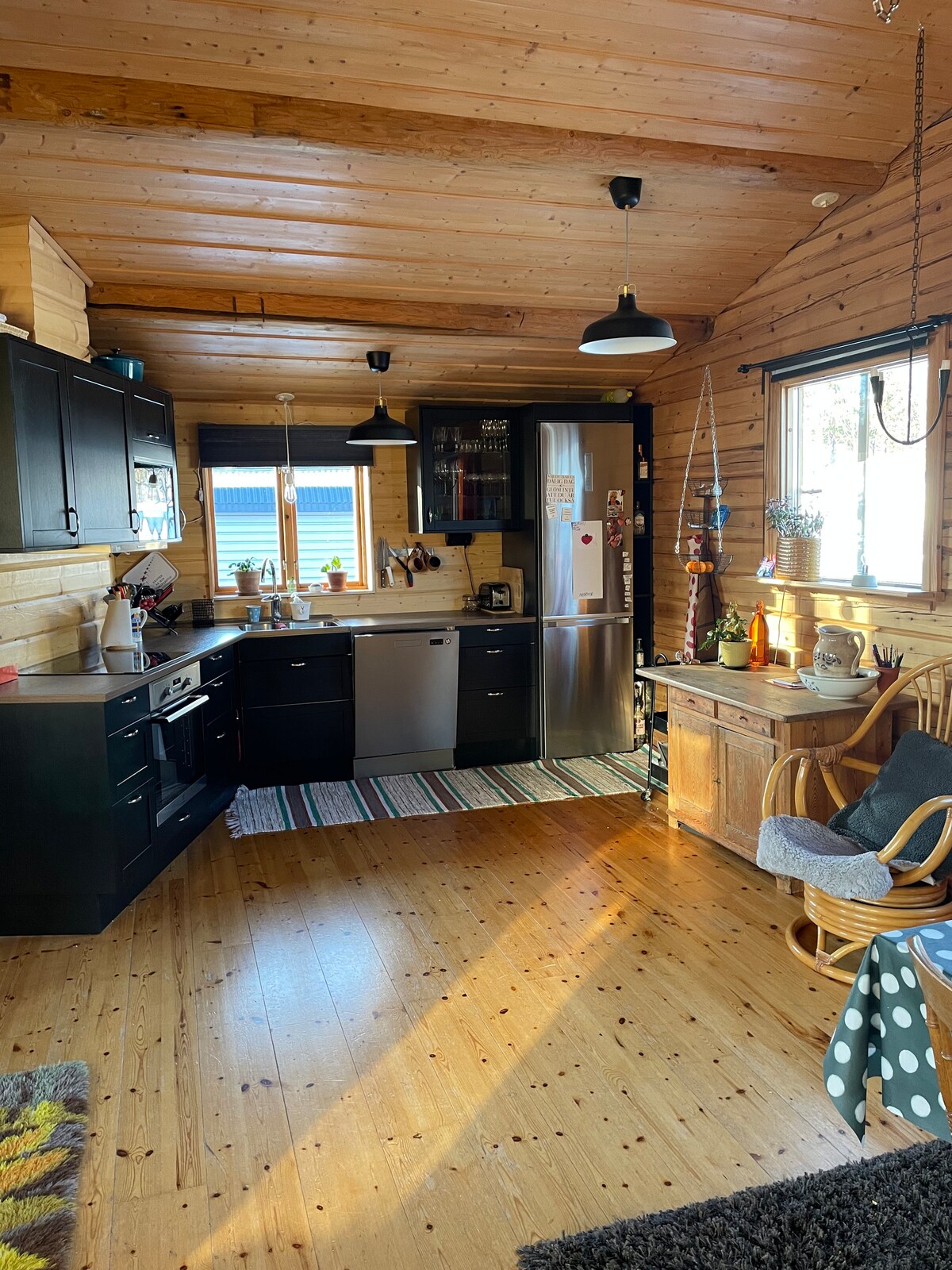 A well-equipped kitchen is featured, characterized by dark cabinets and stainless steel appliances. Natural wood accents create a warm environment, with a wooden dining table and a rocking chair positioned near a window that provides natural light.