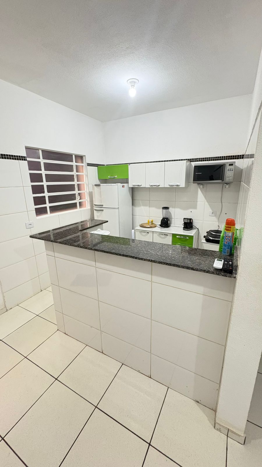A clean kitchen space features white walls and tiled flooring. The modern countertops are complemented by green cabinetry. Appliances, including a refrigerator and microwave, are neatly arranged. Natural light is invited in through a window, enhancing visibility in the cooking area.