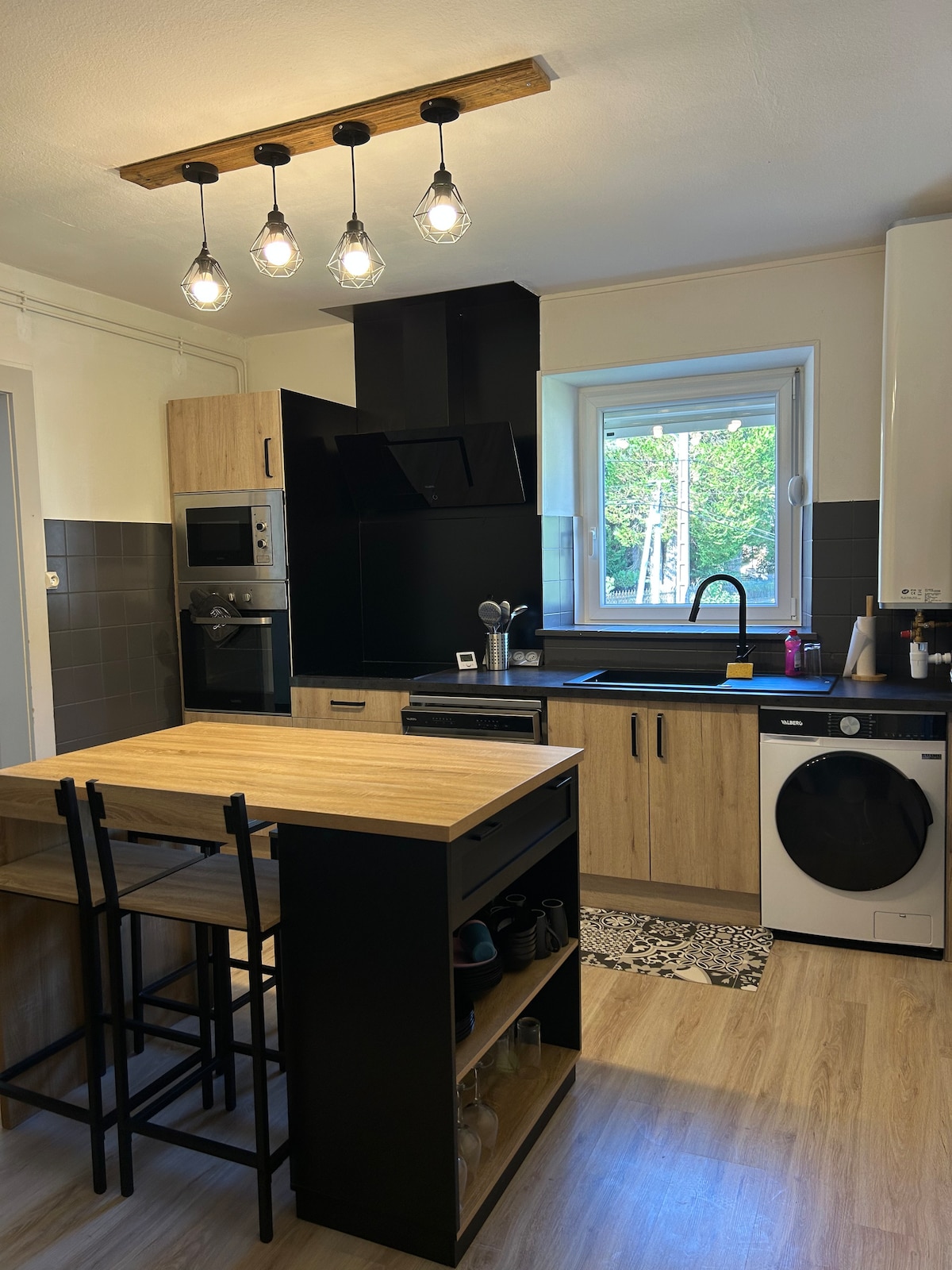 A modern kitchen is presented with wooden cabinetry and dark countertops. A central island with bar stools offers seating for four. Appliances include an oven, microwave, washing machine, and a sleek black sink. Natural light enters through a window, enhancing the functional layout.