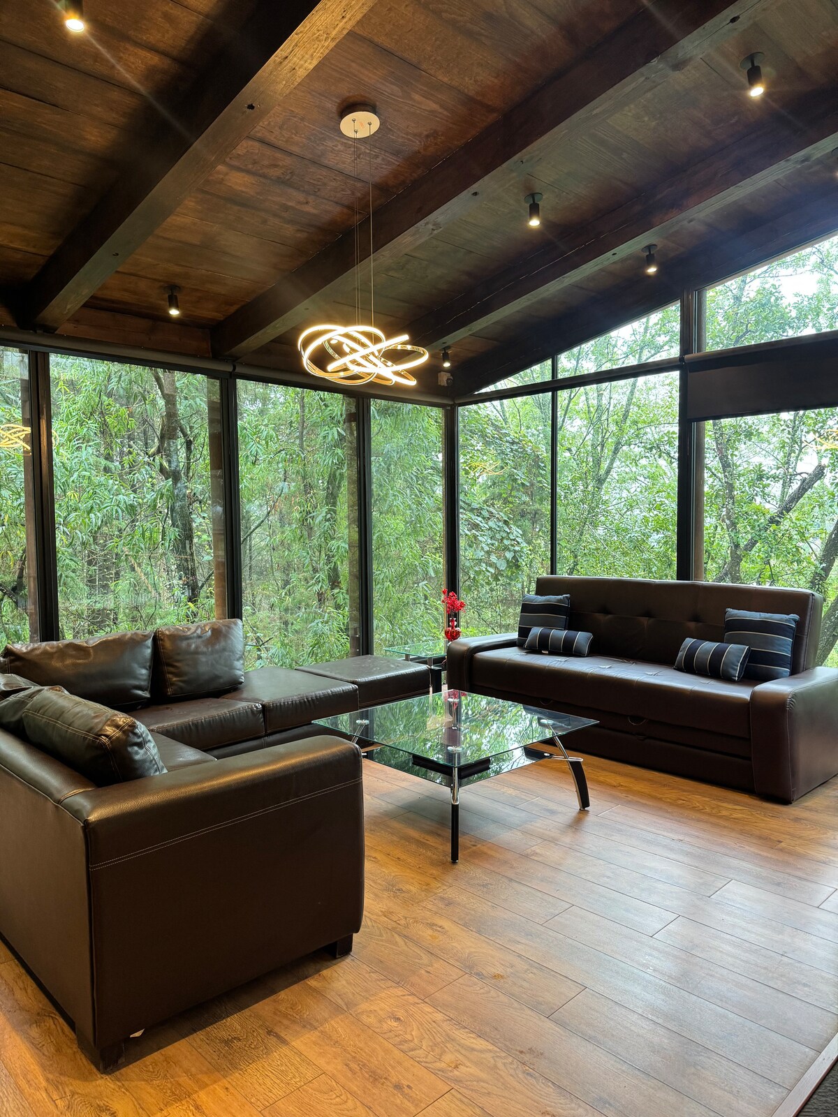 A modern lounge area features dark leather sofas arranged around a glass coffee table. Large windows provide expansive views of the surrounding greenery. A unique light fixture hangs from the ceiling, complementing the wooden beams and creating a contemporary yet serene setting.
