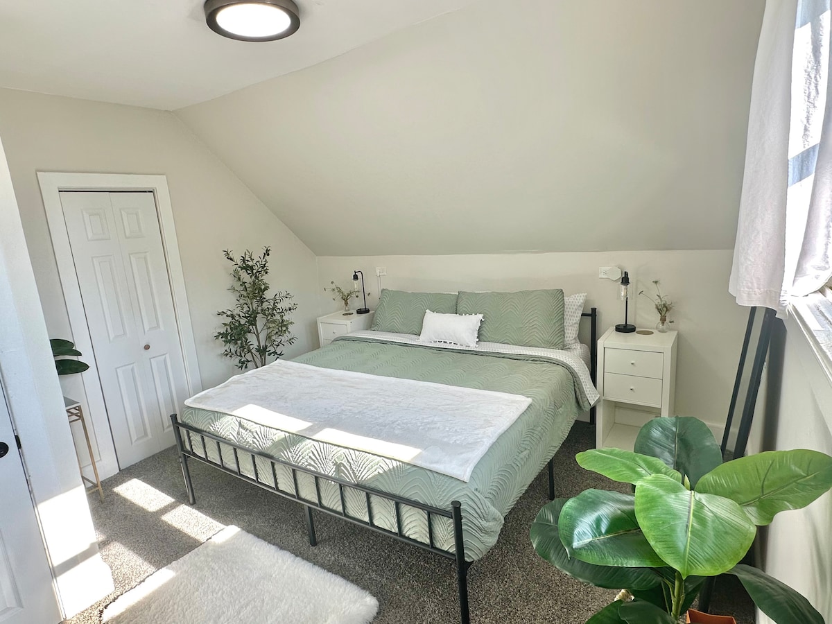 A bright bedroom features a spacious bed dressed in soft green linens, accompanied by white accent pillows. Light pours in through a nearby window, illuminating the room. A small bedside lamp and two nightstands are positioned beside the bed, with greenery placed in the corner.