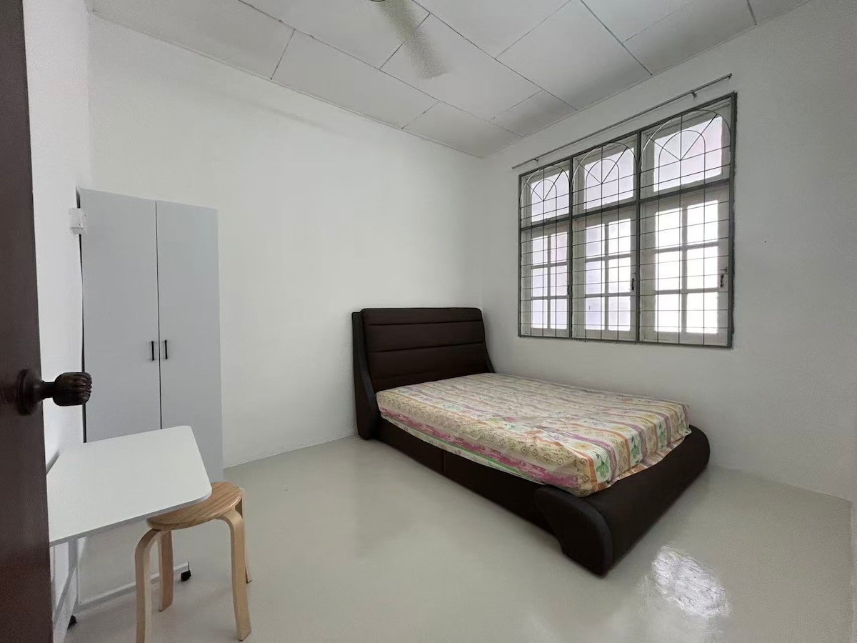 A minimalist bedroom features a spacious bed with a patterned coverlet. A small table and wooden stool are positioned nearby. Large windows with intricate grills allow natural light to fill the room, creating an airy atmosphere against the white walls and glossy flooring.