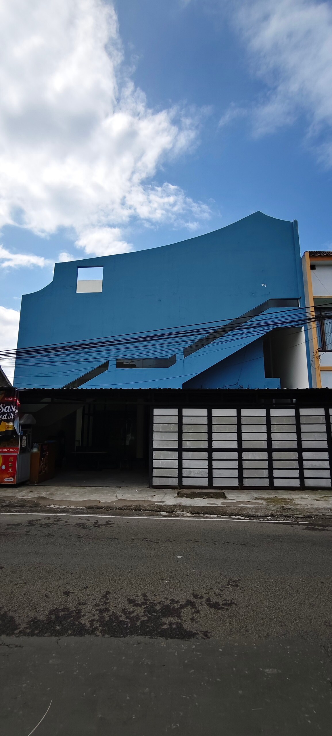 The exterior of a uniquely designed building features a bold blue facade and an angular structure. Subtle details include a clear sky above and overhead power lines. A parking area with a weathered roof is visible in the foreground, enhancing the accessibility of the location.