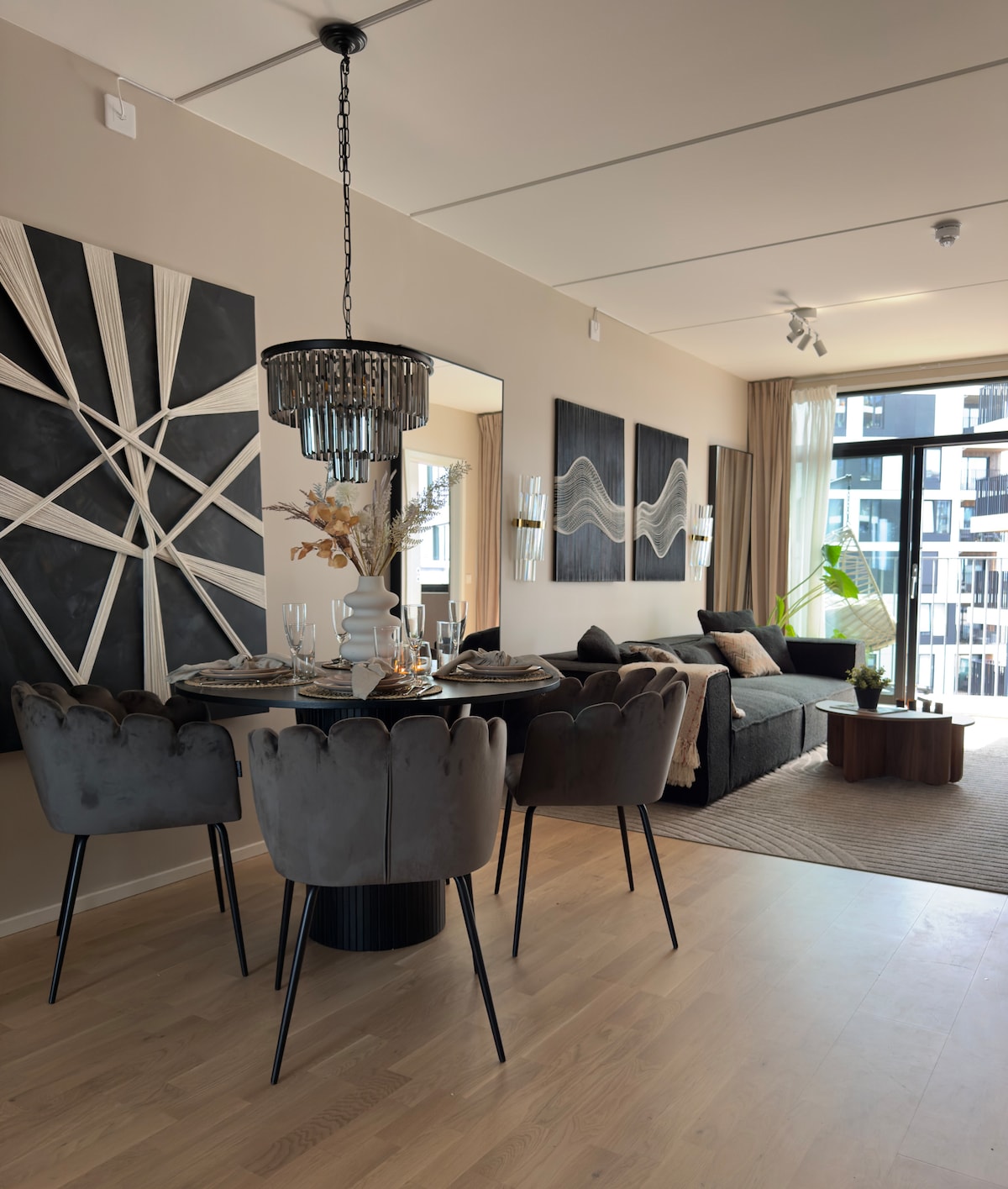 A modern living area is featured, complemented by a stylish dining table with four upholstered chairs. Two striking wall art pieces enhance the ambiance, while a comfortable sofa and decorative elements create a cohesive and welcoming space. Natural light filters through the large windows.