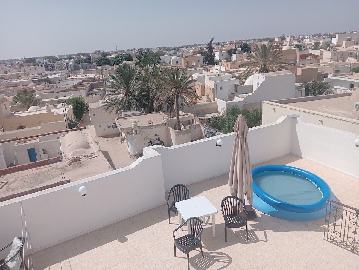 A rooftop terrace is presented with a small inflatable pool, a shaded umbrella, and a table surrounded by four chairs. The view showcases palm trees and the rooftops of Djerbahood, reflecting the local architecture and expansive horizon.
