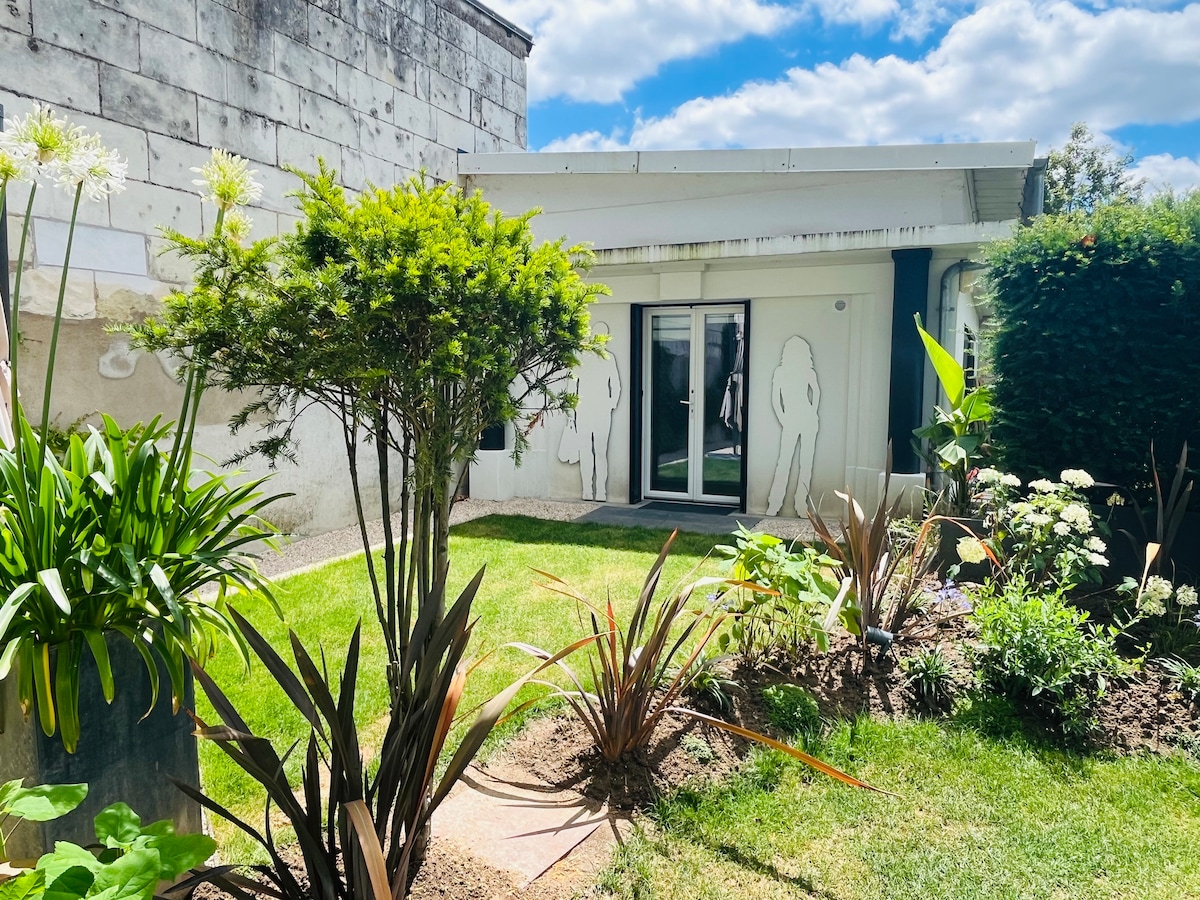 A garden area showcases a well-maintained lawn bordered by various plants and flowers. A sliding door leads to a bright indoor space, complemented by wall art on the building's exterior. Natural light reflects softly against the surrounding greenery, enhancing the tranquil environment.