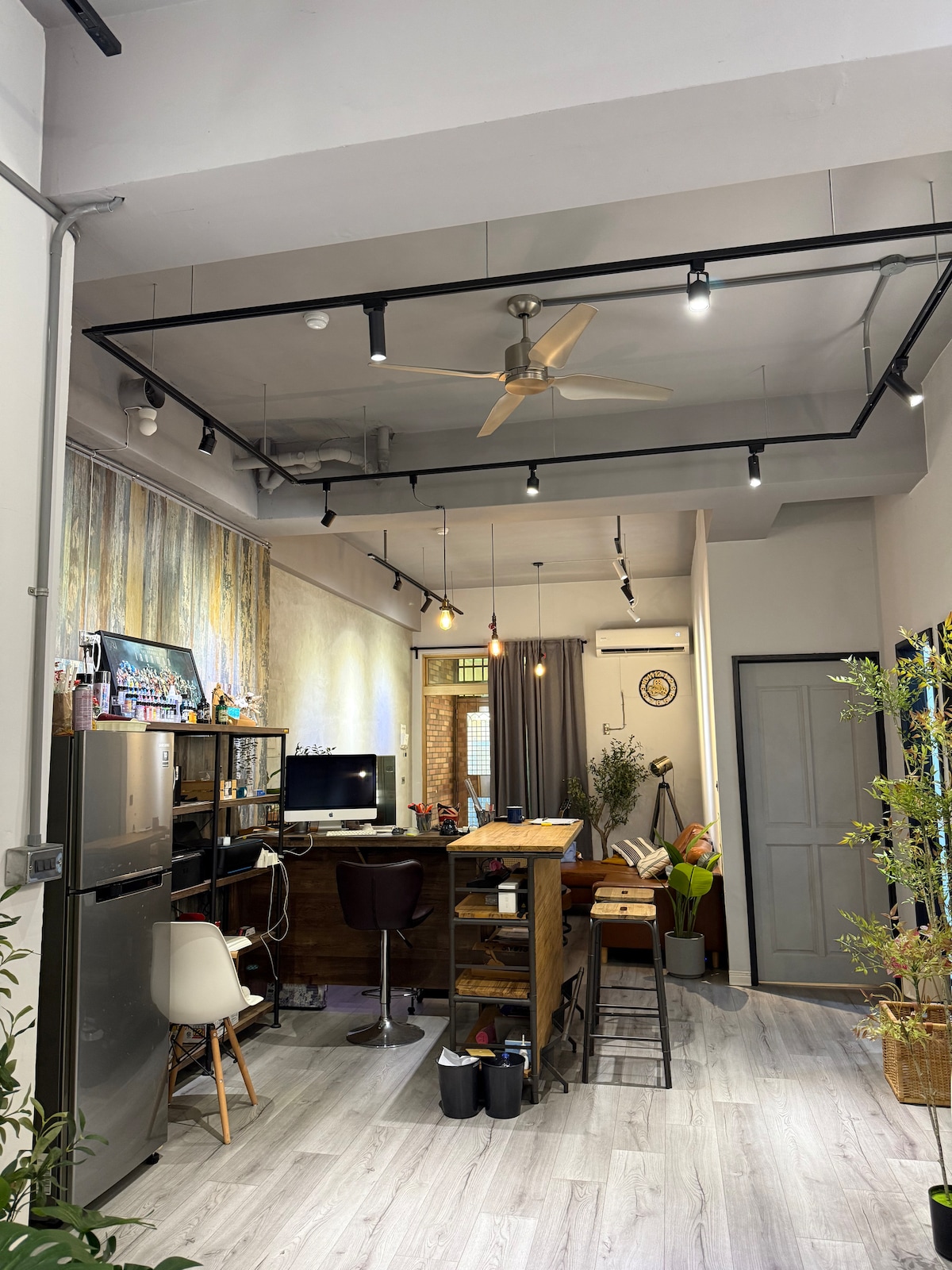 An open living space is showcased, with modern furnishings and a blend of natural light and accent lighting. A kitchen area features a refrigerator and a wooden dining table with bar stools. A ceiling fan enhances airflow in the casually decorated room.