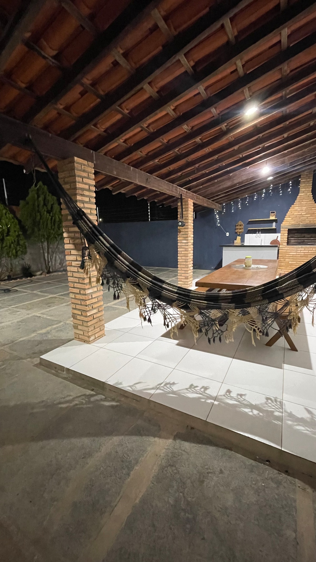 An outdoor barbecue area is highlighted with a rustic wooden table surrounded by wooden benches. A hammock is draped nearby, offering a relaxed seating option. The space is sheltered by a wooden roof supported by brick columns, with a soft light illuminating the area.