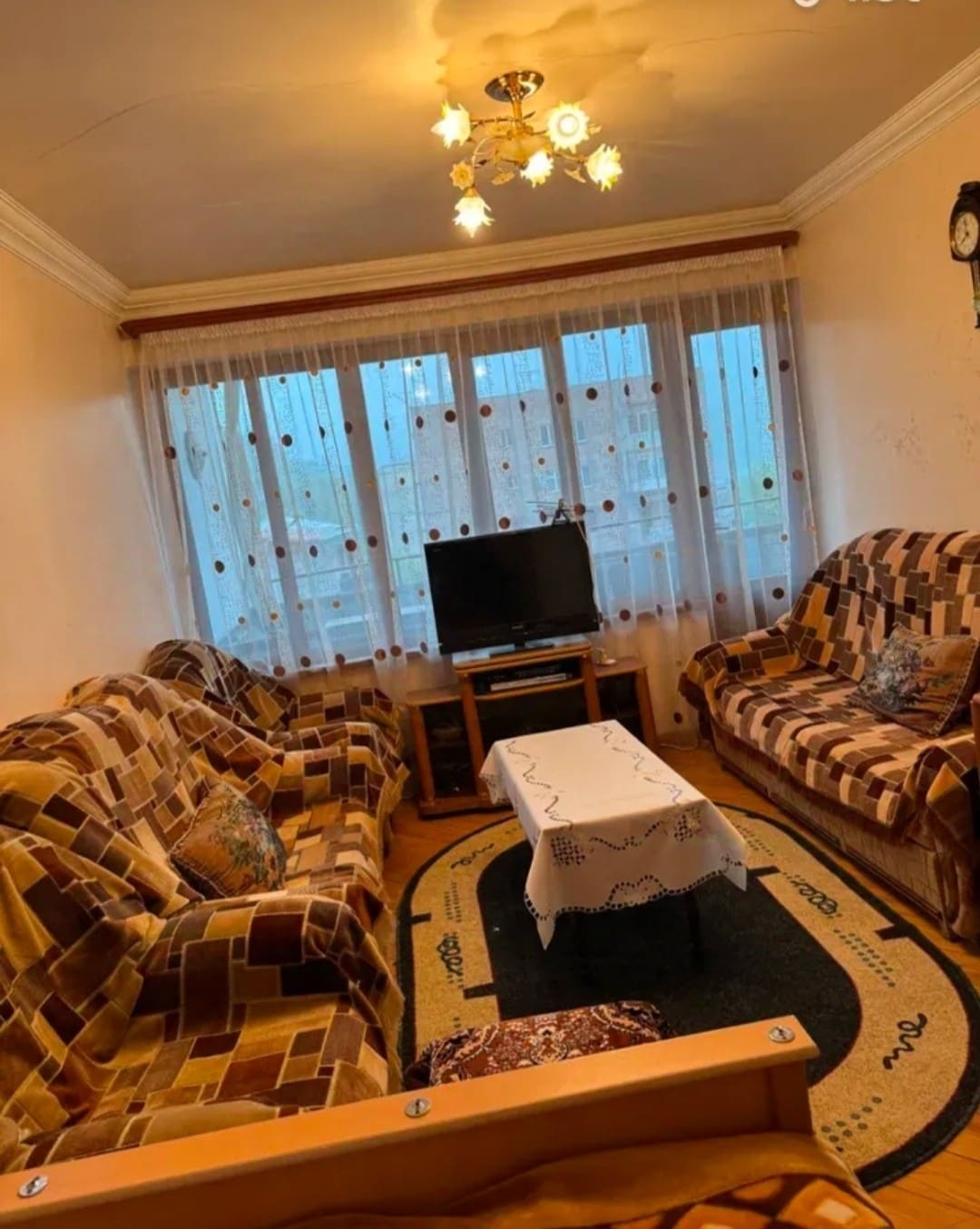 A cozy living area features patterned sofas arranged around a central coffee table. A television is placed on a wooden stand, and large windows with layered curtains allow natural light to illuminate the space. A circular rug adds warmth to the floor.