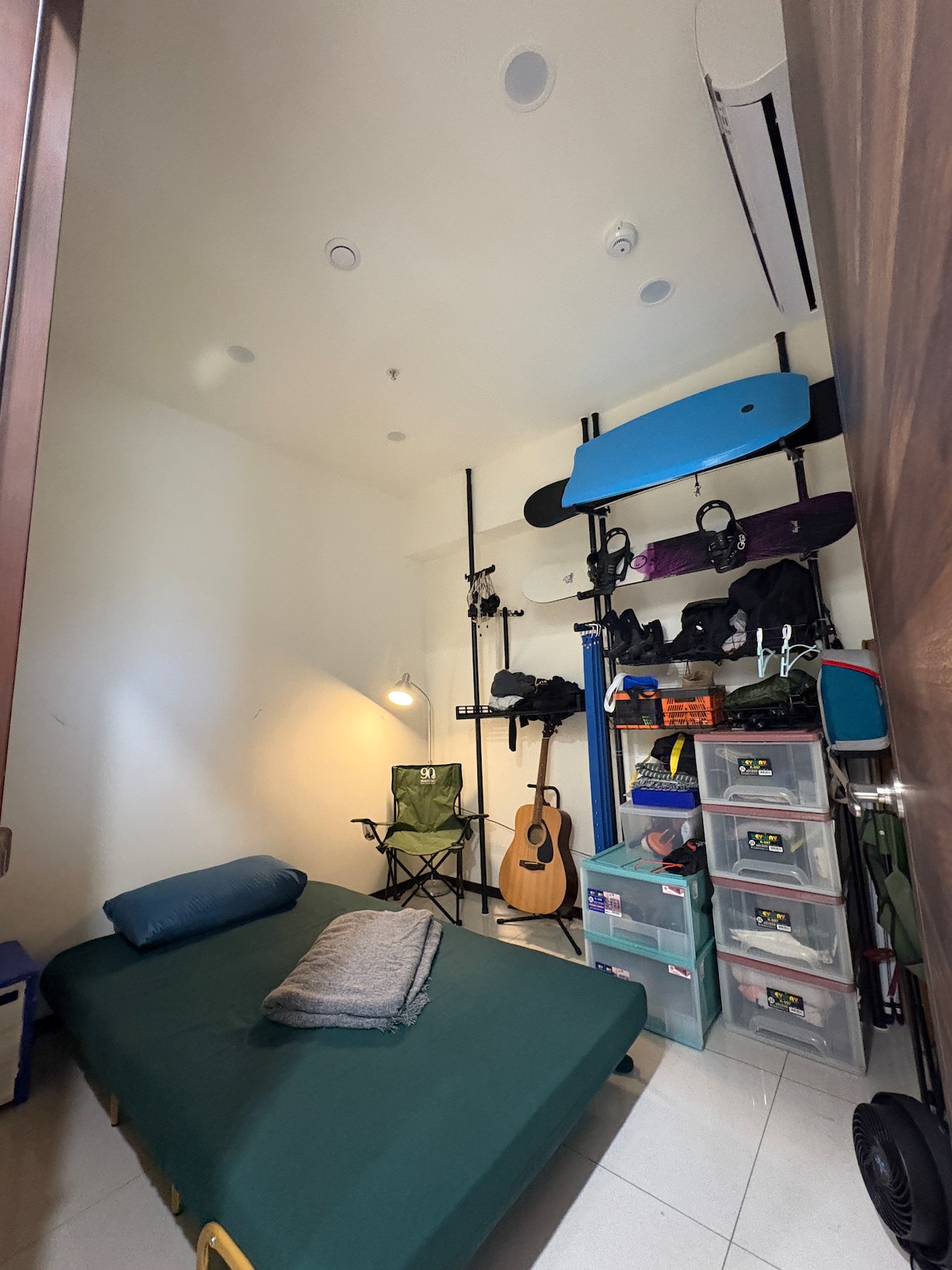 A compact room features a single bed with a folded blanket and a chair in one corner. Equipment for freediving and surfing is neatly organized on the walls, alongside a guitar and storage containers. A window allows natural light into the space.