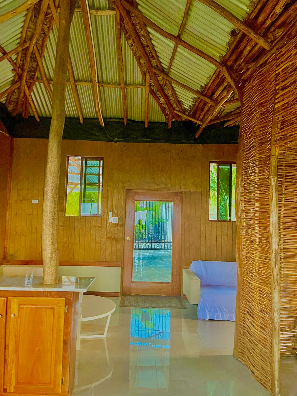 The interior of a cabin features natural wood walls and a high, textured ceiling that allows natural light to filter through. A glass door leads to an outdoor area, while a light blue sofa is positioned near a small, round table. Two windows provide additional illumination.
