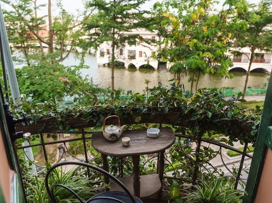 A small balcony is adorned with greenery and a table. A teapot and cup sit ready for use, while a view of a river and lush trees creates a serene outdoor setting, inviting relaxation.