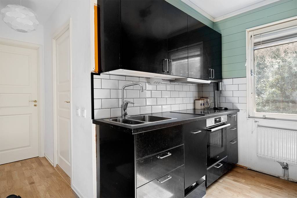 A modern kitchen is shown with sleek black cabinetry, complemented by white tiled backsplashes. Two stainless steel sinks are positioned under a window, allowing natural light. A built-in stove and oven are also visible, with wooden flooring completing the functional space.