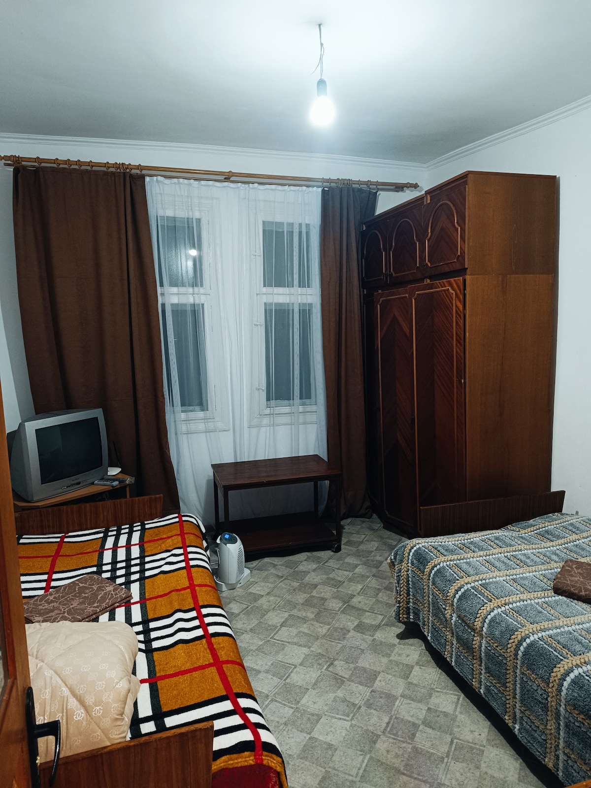 A room features two beds dressed in patterned blankets, with a wooden wardrobe and a small table positioned between them. A television is mounted on a cabinet, and natural light filters through two windows covered by sheer curtains. The floor is tiled with a geometric pattern.