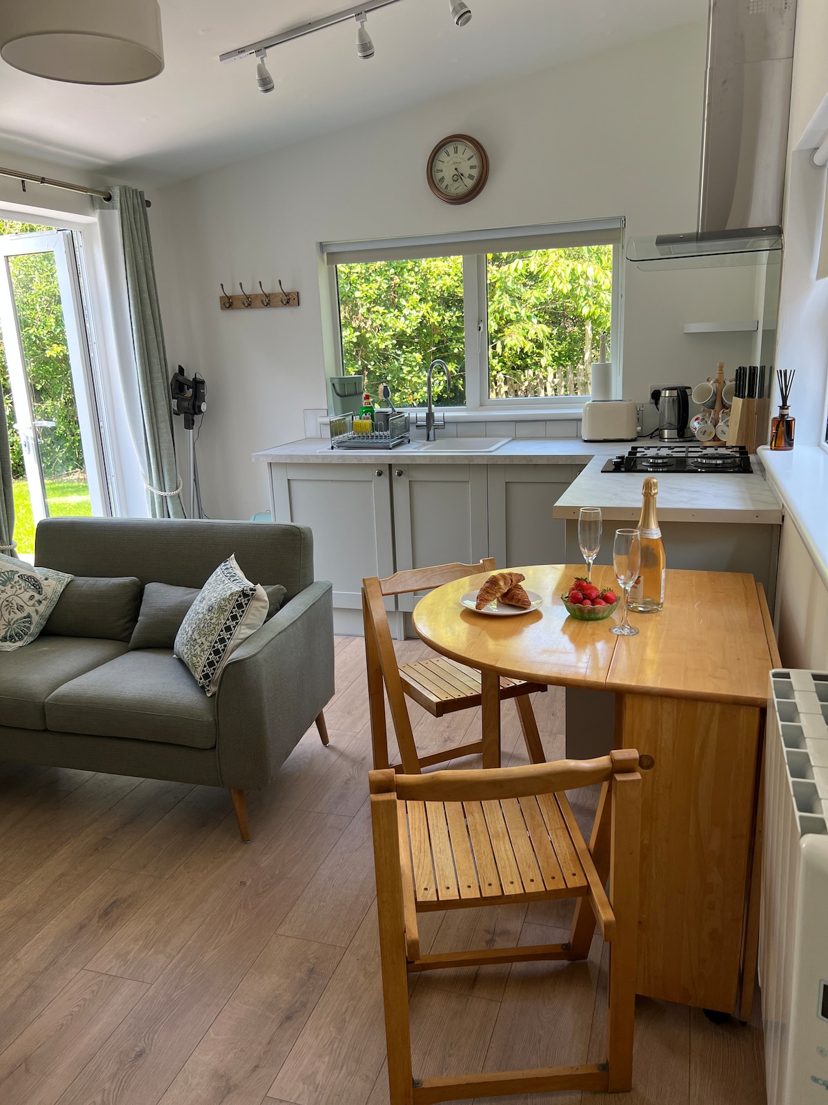 A kitchen area features a wooden table set for two, complete with plates of pastries and fresh strawberries. A grey sofa with patterned cushions sits nearby, and natural light pours in through large windows that overlook the garden.