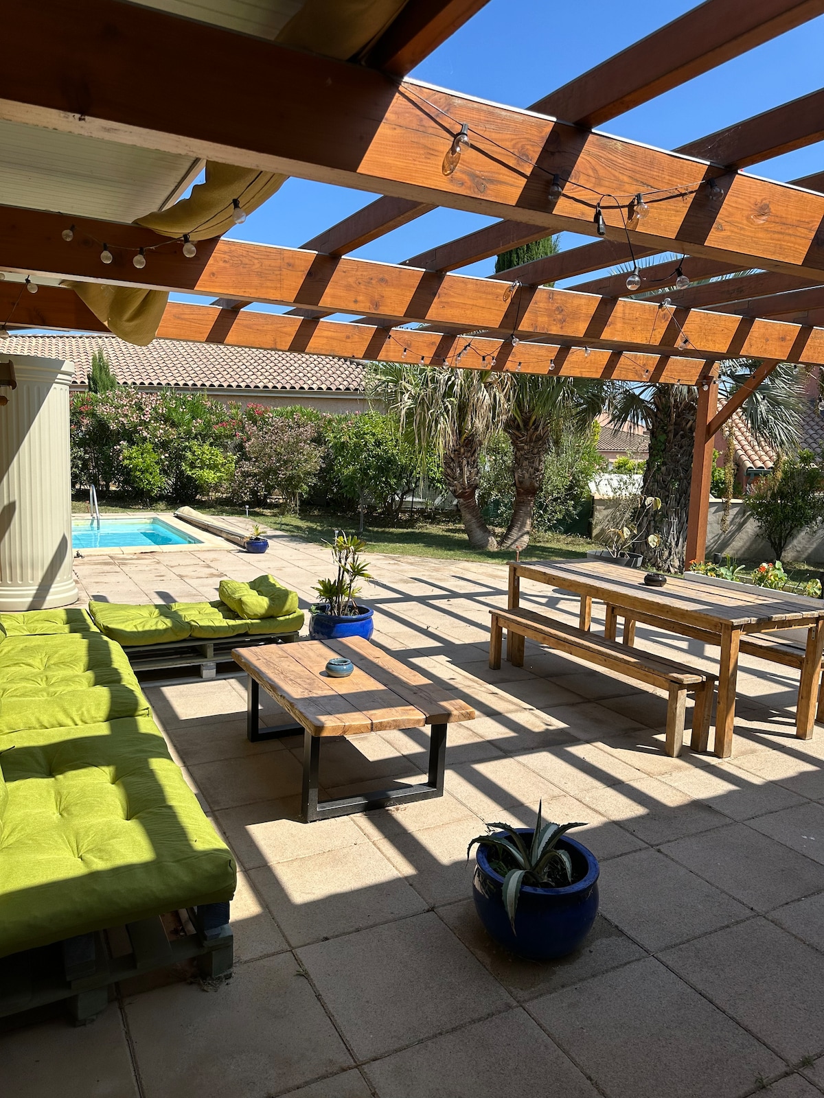 An outdoor terrace is featured, with wooden beams providing structure above. Bright green cushions are arranged on seating areas, while a wooden table stands in the center. The space overlooks a garden with various plants and a private pool in the background, under a clear blue sky.