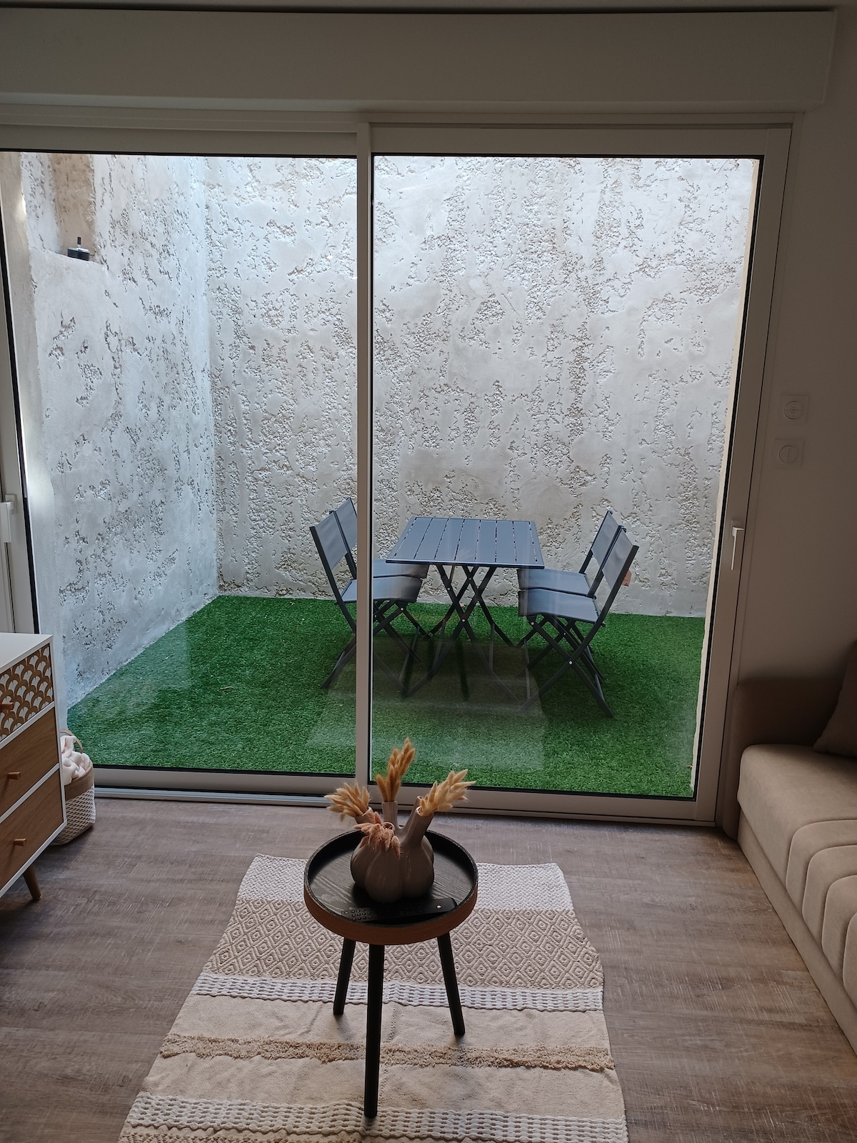 A terrace area is visible through large glass doors, featuring a table surrounded by four chairs on a green carpet-like surface. A textured wall provides a neutral backdrop, while the indoor space includes a small round table with a decorative arrangement.