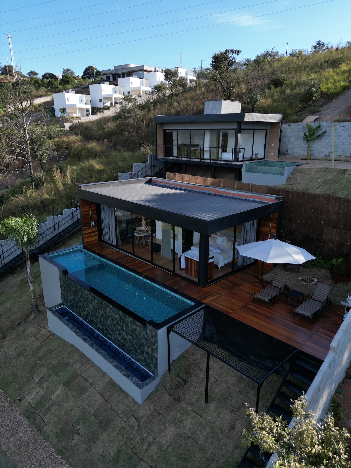 An aerial view captures a modern cabana surrounded by nature, featuring a heated infinity pool that flows into a wooden deck. Sun loungers and an umbrella provide relaxation space, while large glass doors invite natural light into the open living area.