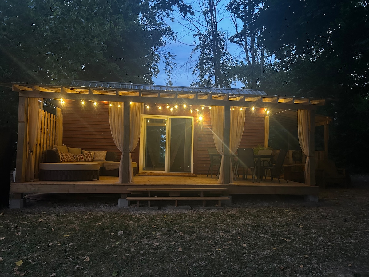 The cabin exterior features a spacious covered porch adorned with soft lighting and flowing curtains. Cozy seating arrangements are present, creating an inviting space for relaxation. Large glass doors provide a view into the cabin, surrounded by lush greenery and a serene atmosphere.