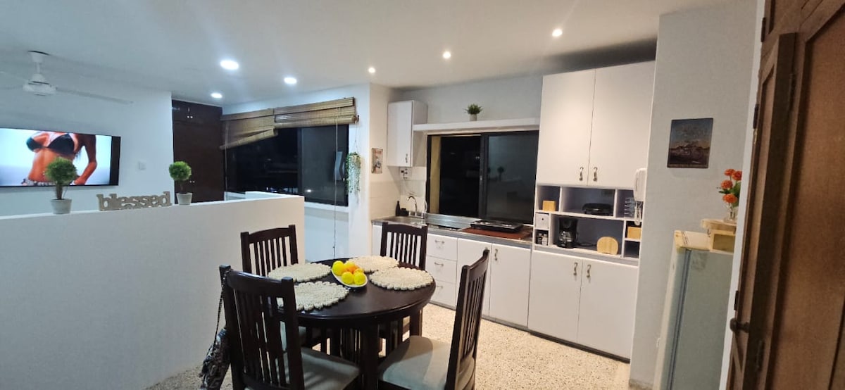 A bright kitchen area is depicted, featuring white cabinetry and a circular dining table. Four wooden chairs surround the table, which is set with placemats and a bowl of fruit. Large windows bring in natural light, enhancing the open and functional layout.