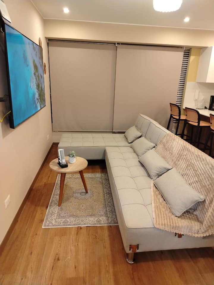 Krásna obývačka s výhľadom na panorámu Limy.
Má veľkú futónovú pohovku so 4 vankúšmi, deku, pohodlnú na odpočinok so 66-palcovou SMART TV s internetom, okrúhly stôl s ovládaním TV a klimatizácie a koberec s béžovými zatemňovacími závesmi.