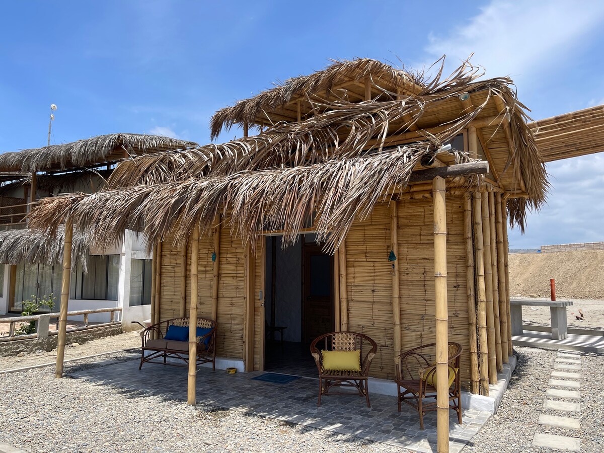 A charming thatched roof cottage is showcased, constructed with bamboo and natural materials. Two woven chairs adorned with colorful cushions are positioned in front, offering a welcoming seating area. The exterior features a sandy pathway leading to an inviting entrance, surrounded by open space.