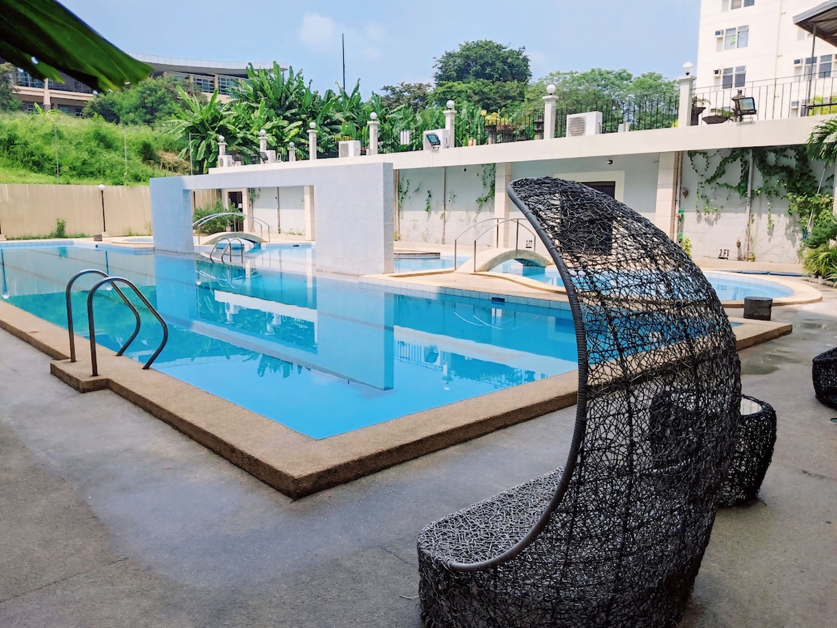 An outdoor pool area is featured, surrounded by greenery. The tranquil water reflects the sky, while unique woven chairs provide seating. Lounge chairs are positioned along the poolside, inviting relaxation in a serene environment.