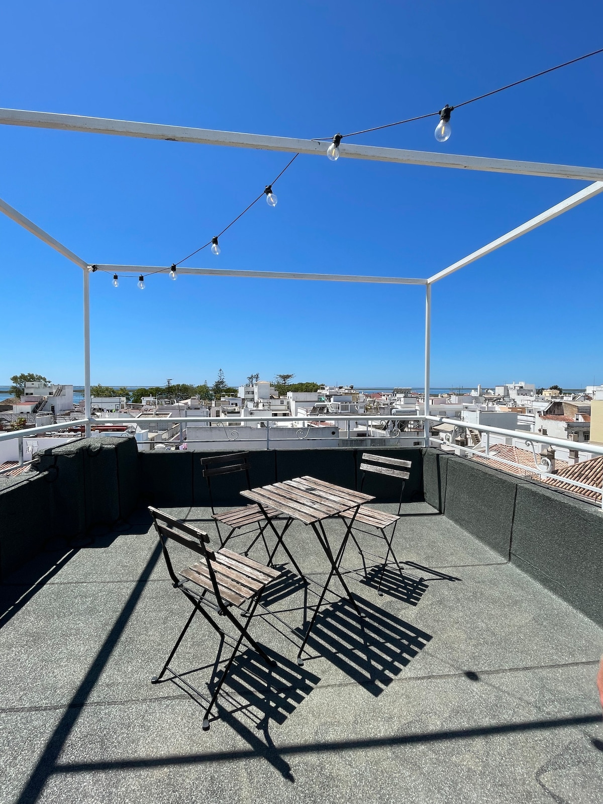 A spacious rooftop terrace is shown, featuring two bistro tables surrounded by four folding chairs. Soft lighting strings are hung above, and the view includes the rooftops of Olhão and hints of greenery in the background under a clear blue sky.