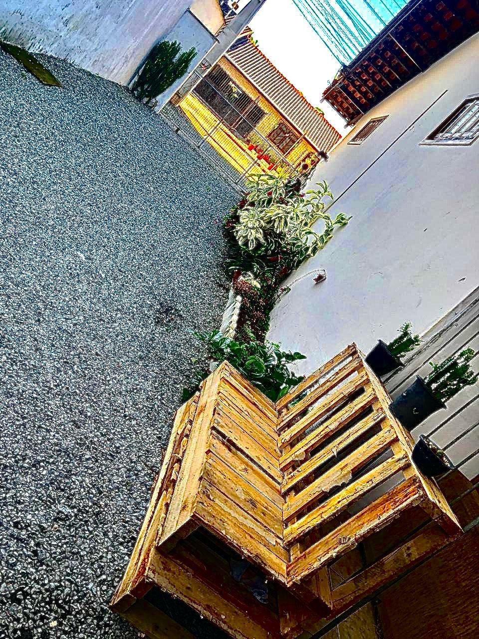 A wooden bench made from pallets sits alongside a pebbled pathway. Lush green plants and flowers line the edges, enhancing the natural ambiance. The pathway leads towards a well-lit area, providing a sense of openness and tranquility.