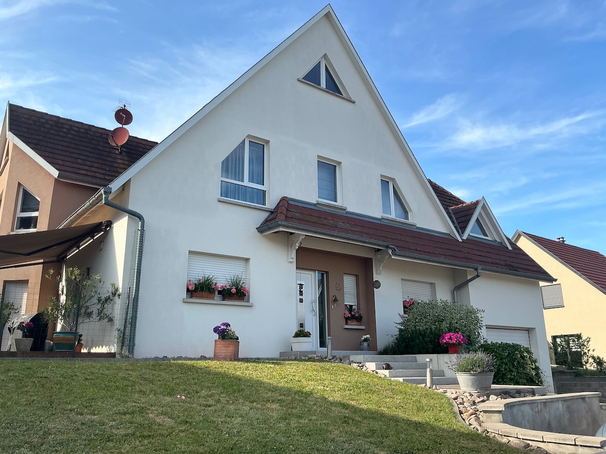 A modern, two-story building is depicted with a sloped roof and multiple windows. Flower boxes adorn the front, adding touches of color. A well-kept grassy area leads to the entrance, which is highlighted by steps and planters. Blue skies provide a serene backdrop.