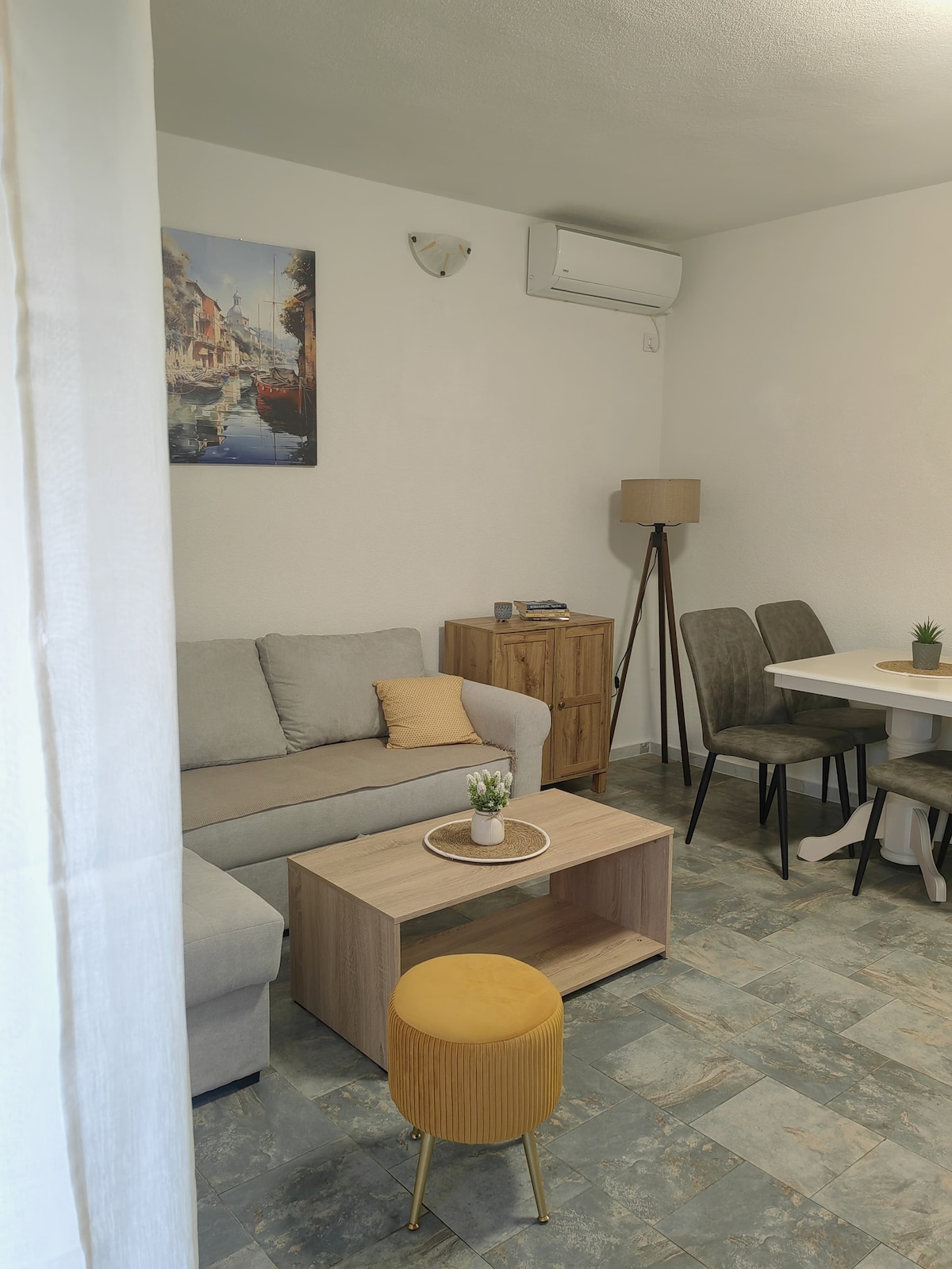 A light-filled living area features a comfortable sofa, a small wooden coffee table, and a yellow ottoman. A dining table with four chairs is positioned nearby, complemented by a tall lamp and a piece of wall art depicting a seaside scene.