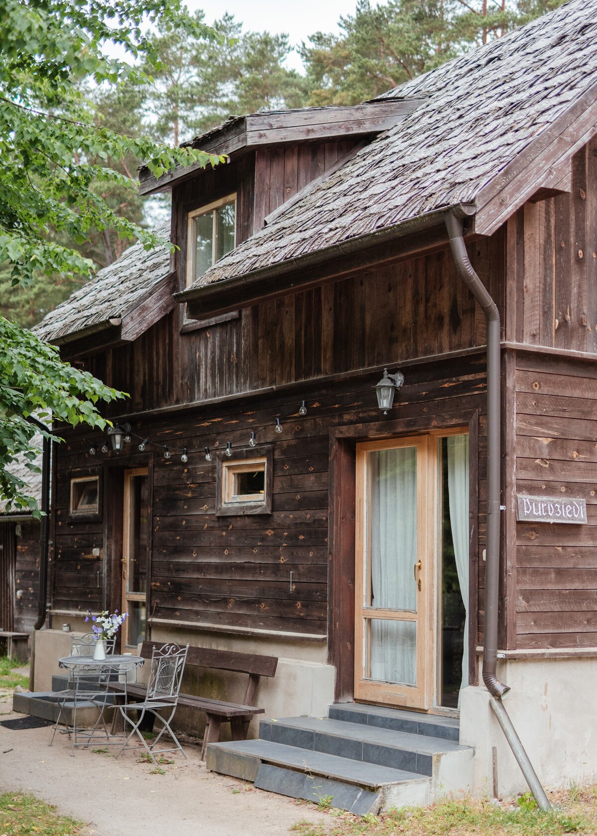 A rustic wooden cabin is framed by lush green trees, featuring a cozy outdoor seating area with metal chairs and a small table. Large windows allow natural light to fill the interior, showcasing a welcoming entrance with a wooden door.