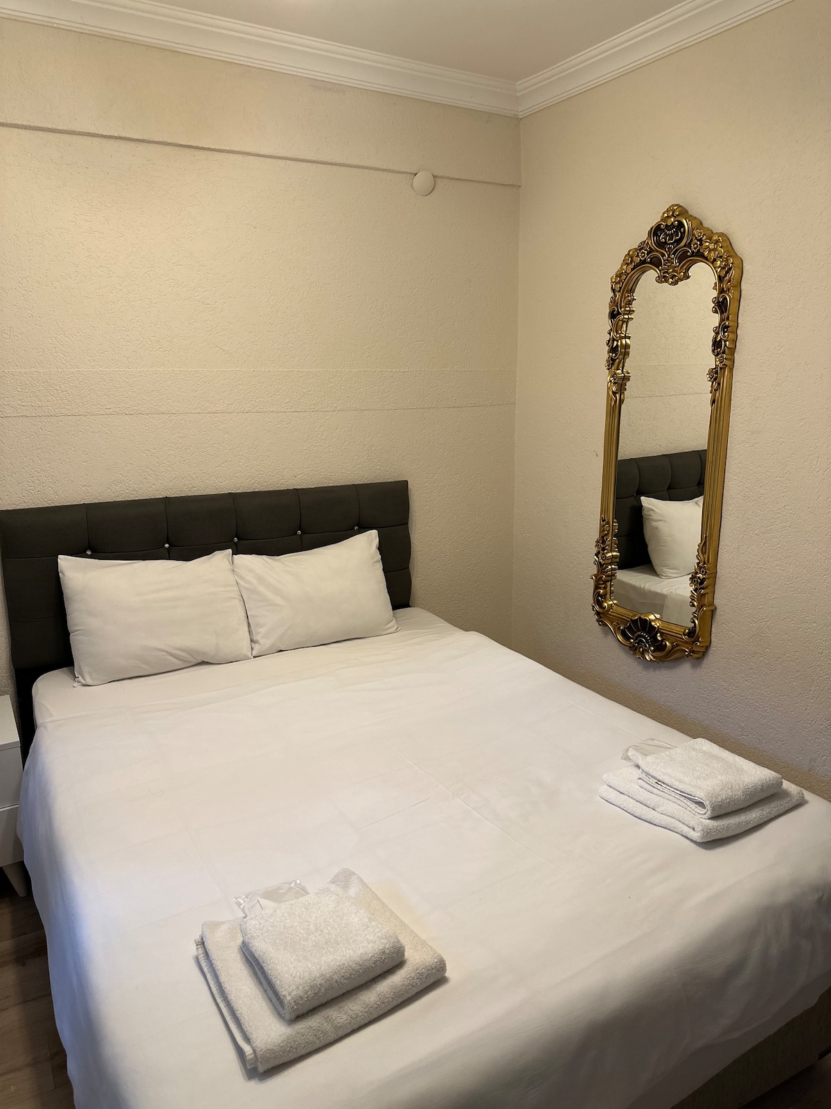 A neatly arranged sleeping area features a spacious bed with crisp white linens and two folded towels placed at the foot. A decorative gold-framed mirror reflects the room, enhancing the light and airy ambiance. The soft, neutral walls contribute to a calming environment.