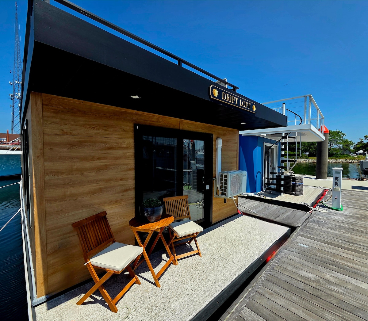 The exterior of the modern houseboat features a wooden façade and large windows. A small patio area includes two wooden chairs with white cushions, positioned on a textured surface beside the water. The setting is bright under a clear blue sky.