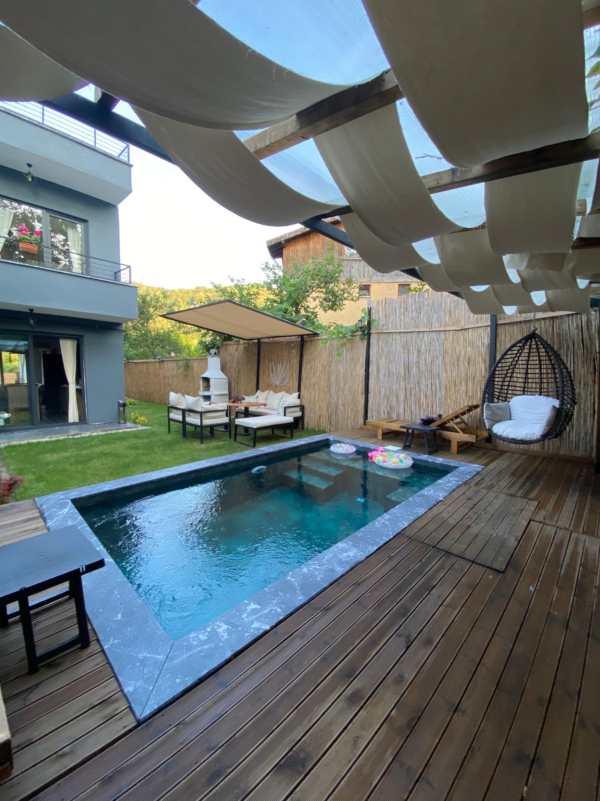 An outdoor area features a compact pool surrounded by wood decking, with lounge chairs positioned nearby. A shaded seating area is visible, along with a hanging chair and decorative elements such as floating pool toys. The surrounding greenery adds a relaxing touch.