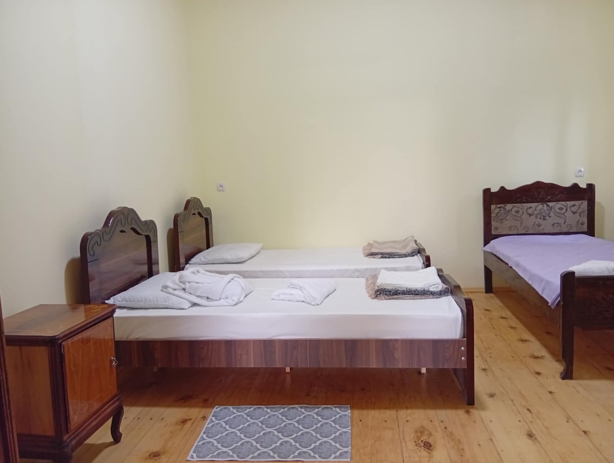 A room featuring two beds with simple wooden frames, one covered with a plain purple blanket. A bedside table is positioned alongside one bed, while towels are neatly arranged on the beds. Light-colored walls and a wooden floor create a bright, open feel.