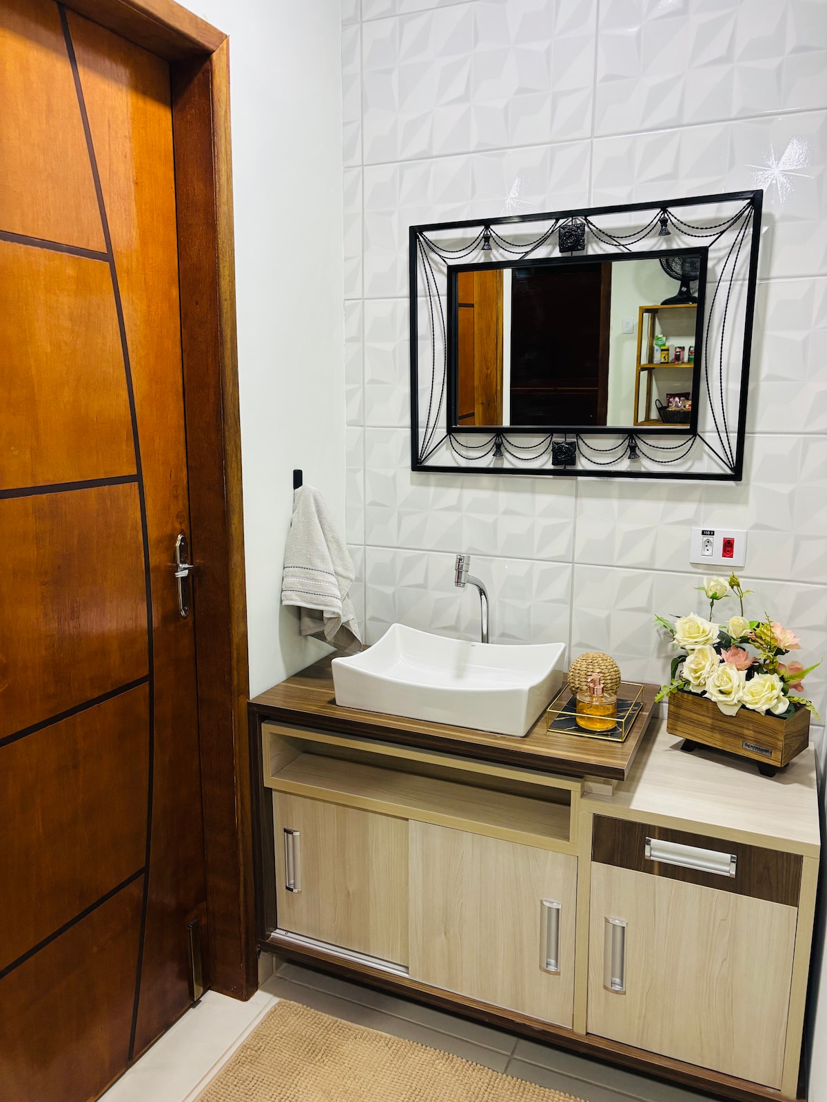 A modern bathroom features a wooden door leading to the space. A sleek sink is positioned on a countertop with storage below. Decorative flowers are placed on the counter, complemented by a stylish wall mirror framed in black. Light-colored tiled walls enhance the room's design.