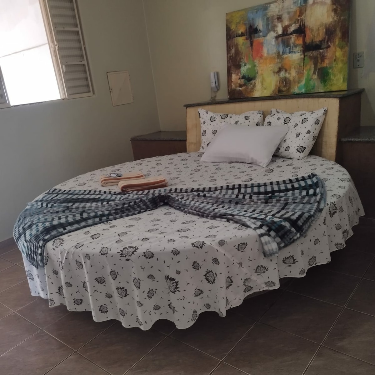 A round bed with a floral-patterned bedspread is positioned centrally in the room. A light-colored blanket is casually draped over the bed, complemented by two white pillows. A piece of abstract wall art adds color above the headboard, while natural light enters through a nearby window.
