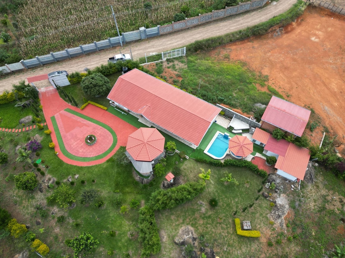 An aerial view captures the private property featuring a well-maintained garden, vibrant green areas, and a swimming pool. The buildings are outlined by a red-tiled roof and pathways, framed by lush foliage and grass, creating a serene environment ideal for relaxation.
