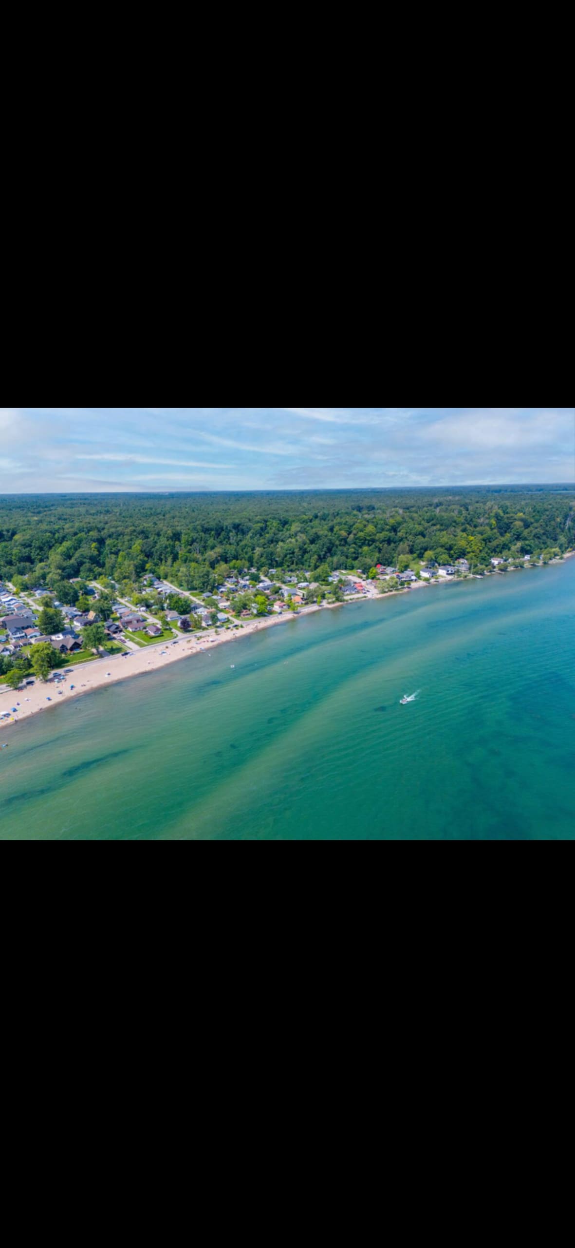 Aerial view of a coastline featuring a sandy beach that gently curves along the shore. Crystal-clear water transitions from turquoise to deep blue, with a boat visible on the water. Lush greenery borders the beach, creating a serene natural backdrop.