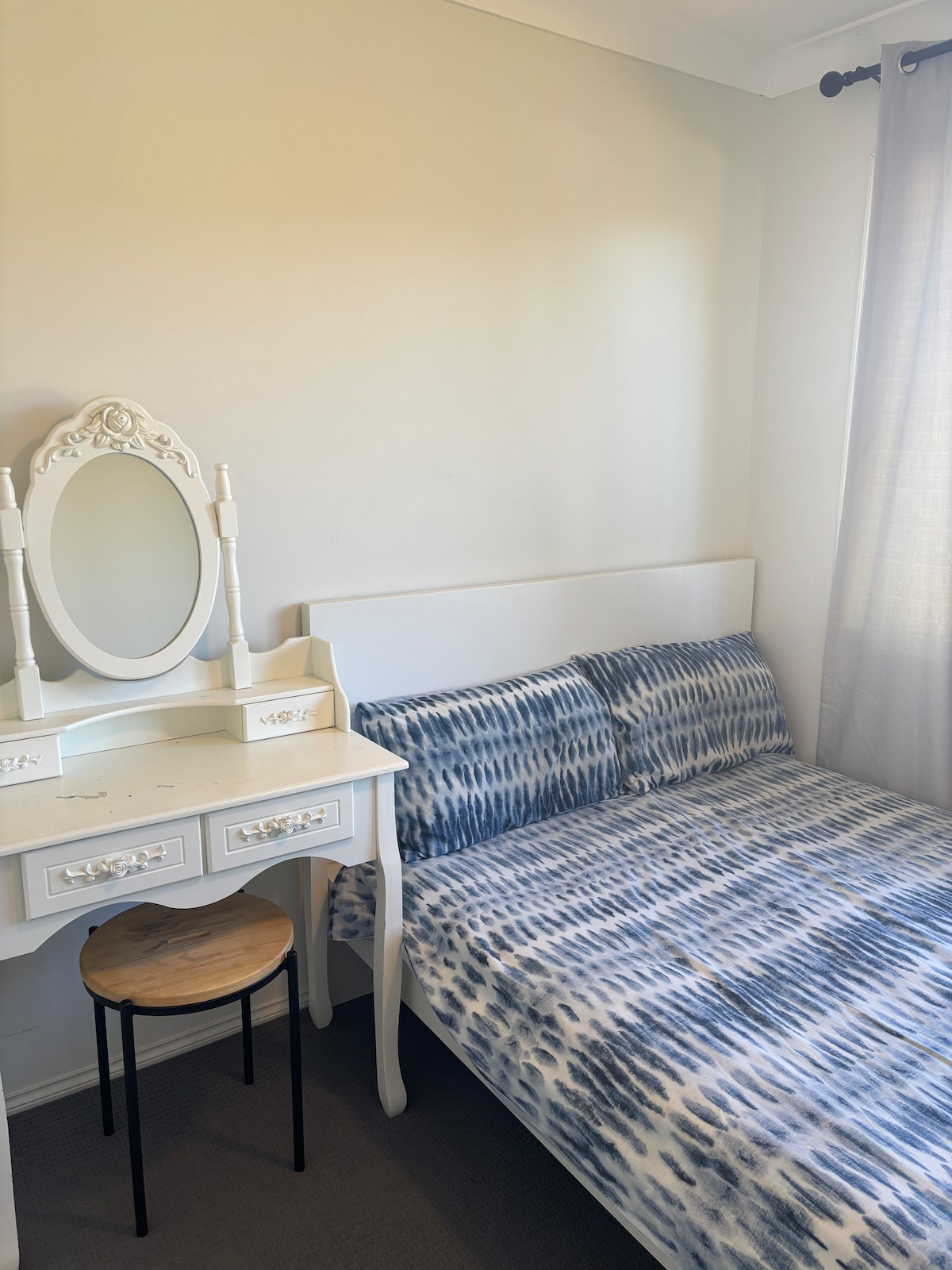 A light-filled bedroom features a bed dressed in blue and white patterned bedding. A small wooden stool is placed beside the bed. A white vanity with a round mirror adds elegance to the space, while soft curtains gently filter natural light.