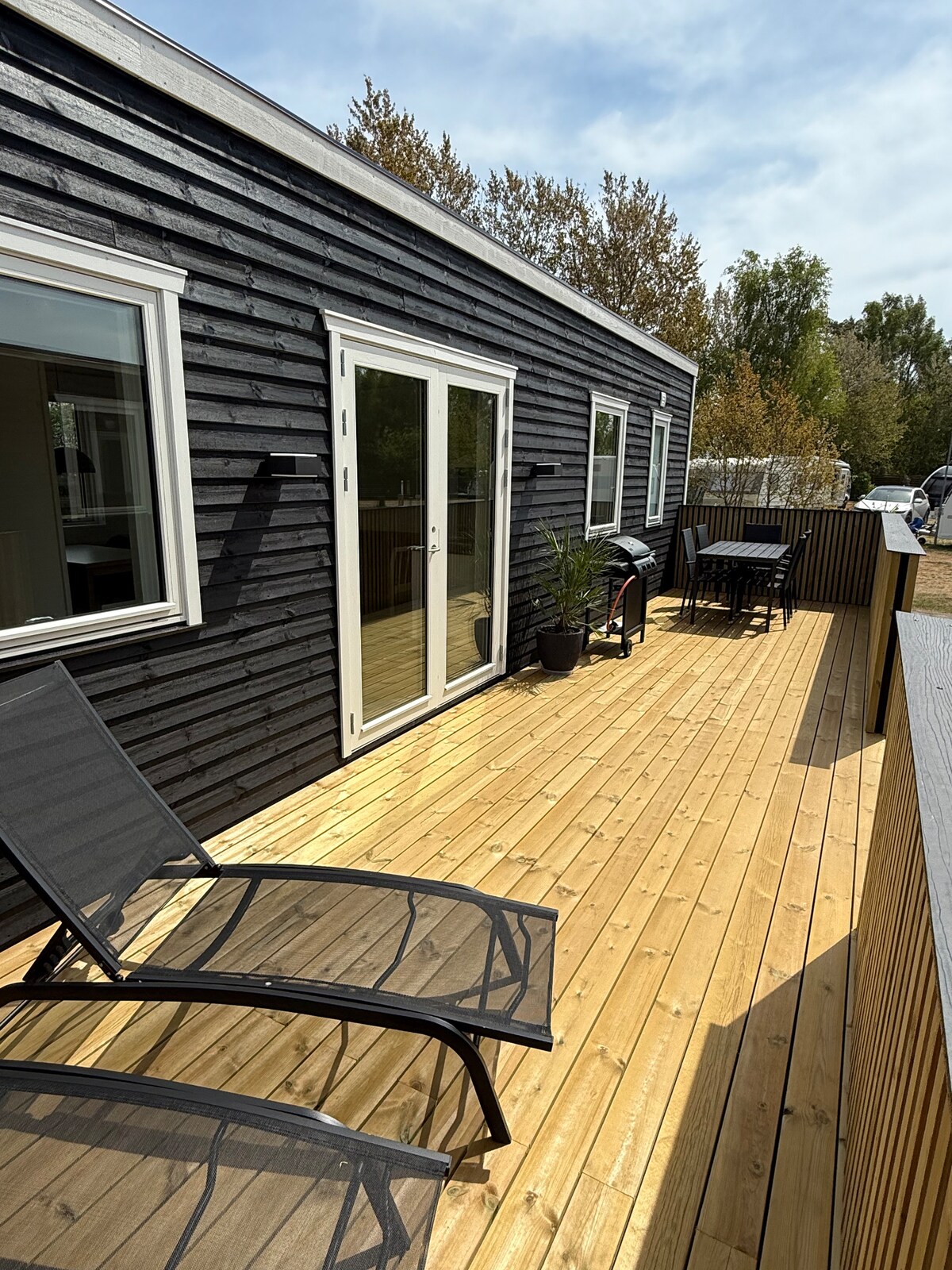 A spacious wooden deck is featured, adorned with two lounge chairs and a dining set for four. Large glass doors open into the living area, allowing natural light to enhance the space. Surrounding trees add a touch of greenery to the serene setting.