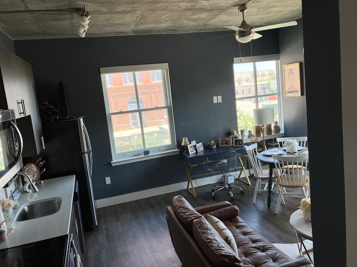 A modern kitchen and dining area occupy an open floor plan. Dark walls frame large windows that provide natural light. A cozy living space includes a leather sofa, while a round dining table is complemented by white chairs, creating an inviting setting.