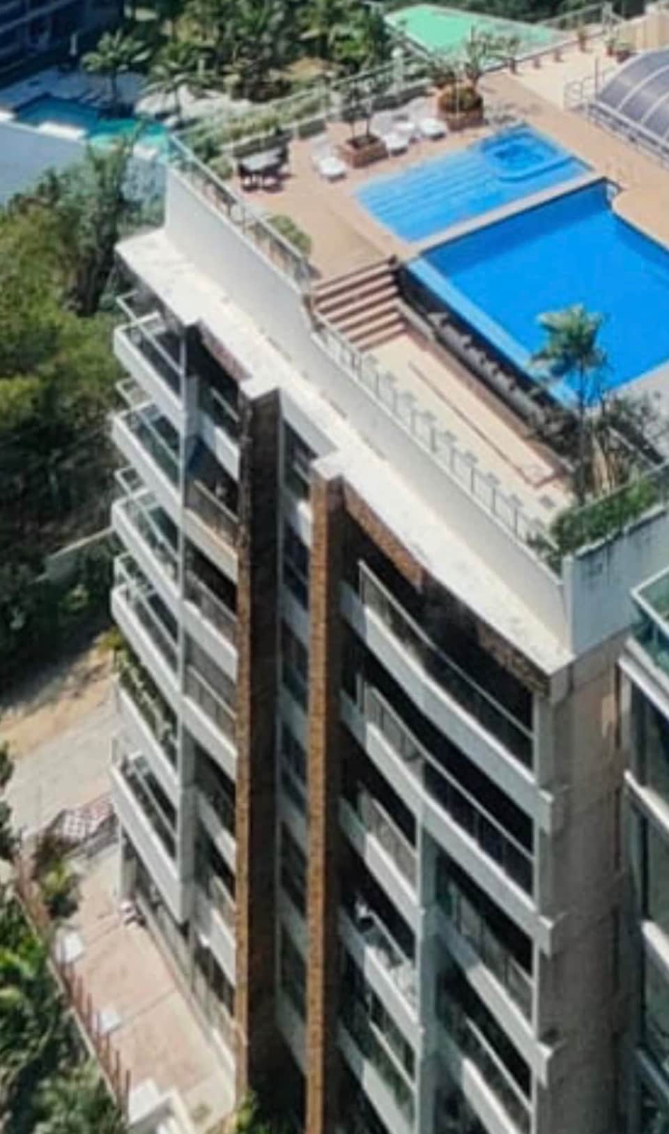 The image displays a modern building with multiple floors, topped with a large terrace featuring a swimming pool. Lounge chairs and greenery are visible on the terrace, while the surrounding area showcases a blend of lush vegetation and urban landscape.