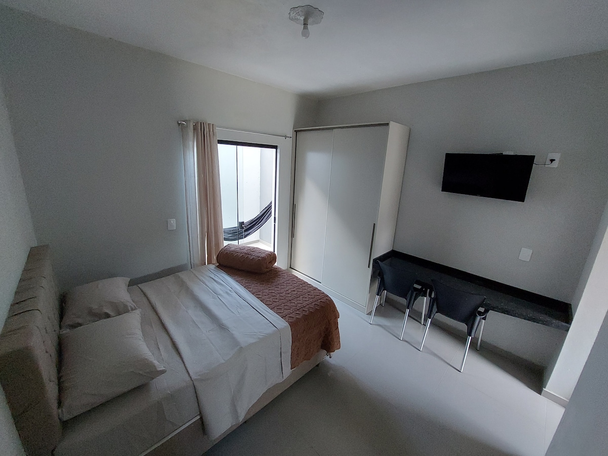 A serene bedroom features a spacious bed with neutral-toned bedding. A modern television is mounted on the wall opposite a sleek desk with two chairs. Natural light enters through a sliding door, which opens to a private outdoor area, enhancing the tranquil ambiance of the room.