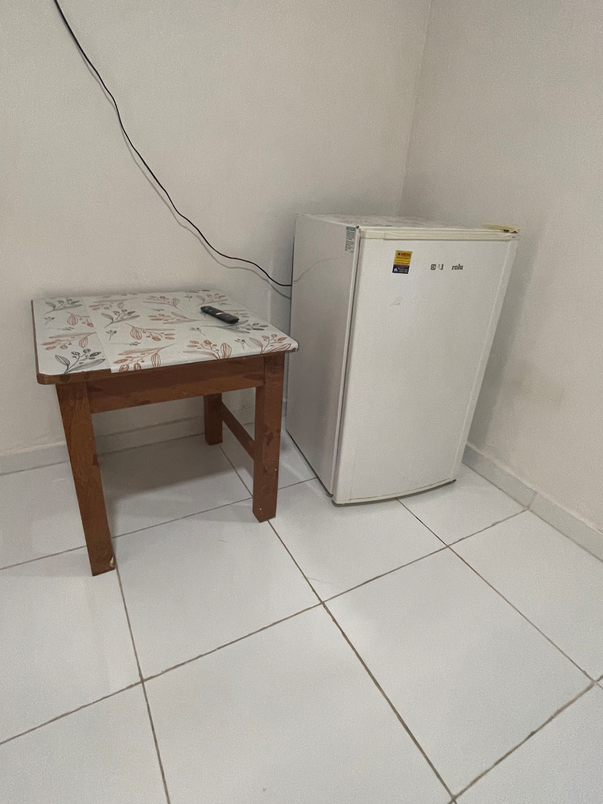 A wooden table with a decorative top is positioned beside a compact white refrigerator. The floor is tiled in a light color, and the walls are painted in a neutral shade, contributing to a clean and functional space.
